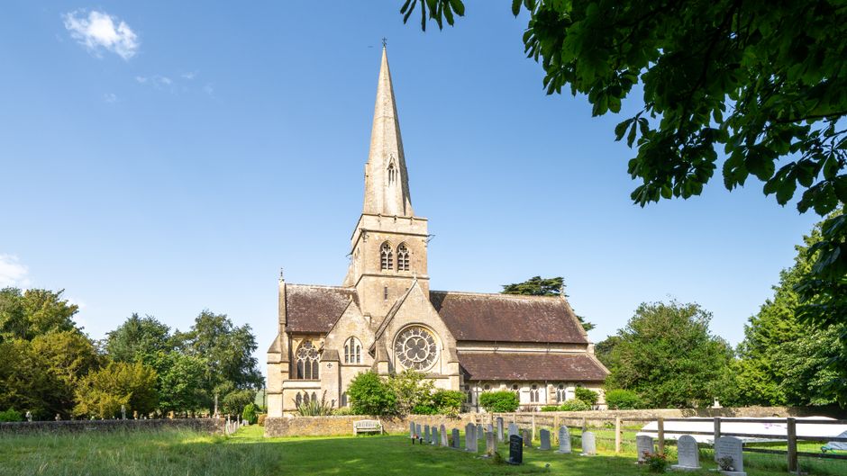 A historic stone church with a tall, pointed spire dominates the scene, situated in a lush, green cemetery with neatly arranged white and gray headstones along the grass. The church features a large circular stained glass window on the front facade, arched windows, and a pitched roof with dark shingles. The surrounding environment includes well-maintained trees and shrubs, some with dense foliage, framing the image on either side. The church is illuminated by natural daylight, with a clear blue sky overhead, and shadows cast on the building's stone exterior highlighting its architectural details. In the foreground, a low stone wall separates the cemetery grounds from the open grassy area, which is part of a tranquil outdoor setting. This scene, captured during daytime, closely relates to the context of managing outdoor spaces, including the potential for rubbish removal and site clearance services that Waste Collection Sutton may offer for such heritage sites or private properties, emphasizing the importance of maintaining clean, accessible outdoor environments.