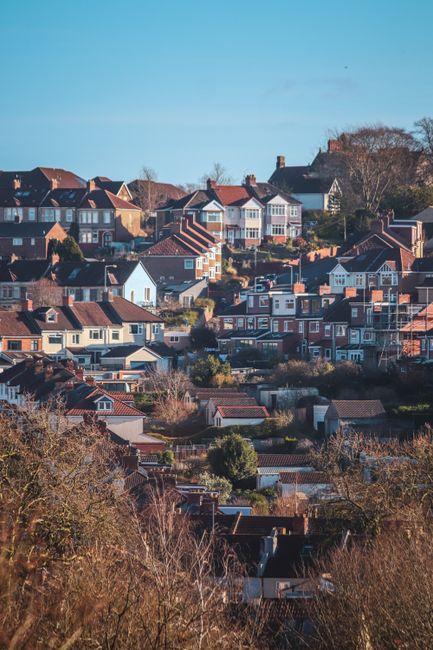 A panoramic view of a residential hillside area during daylight, showing a densely packed collection of houses and bungalows with brick and painted facades in shades of red, white, and cream. The rooftops are predominantly tiled, with some featuring dormer windows and chimneys. The property sizes vary, with some dwellings closely aligned while others are separated by small gardens or pathways. The scene includes mature trees and shrubs interspersed among the homes, contributing touches of greenery against the urban backdrop. The sky above is clear and blue, casting natural light across the landscape, highlighting the textures of the roofs, walls, and foliage. The overall image captures a quiet, suburban environment typical of a stable residential community, which might benefit from private waste handling or alternative rubbish collection services by Waste Collection Sutton, especially in relation to on-site clearance or bespoke rubbish removal needs specific to this type of densely populated area.