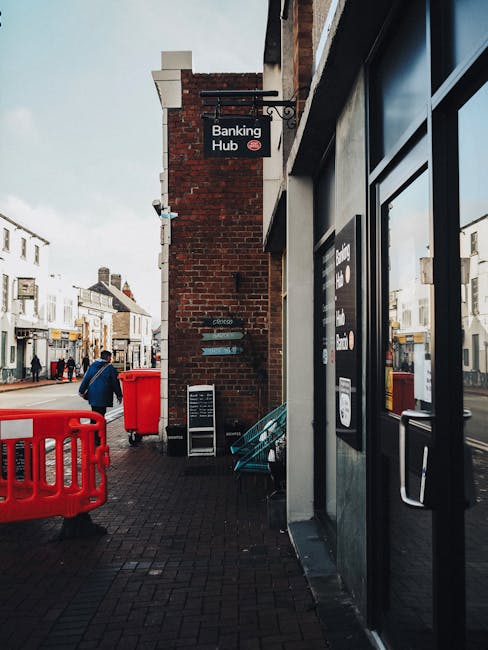 The image shows a street scene outside a building with a brick exterior, featuring a black sign reading 'Banking Hub' mounted above the entrance, which is flanked by dark-framed glass doors. Adjacent to the entrance, there is a large window with a sign indicating banking services. In the foreground, a bright red plastic barrier and a matching red waste bin are positioned on the paved sidewalk. To the left, a person dressed in a blue coat and carrying a backpack walks past, while in the background, several pedestrians are visible on the bustling street lined with white terraced shops and buildings. The environment appears overcast, with diffused natural light illuminating the scene. On the pavement, near the building, there are a few stacked turquoise chairs and a small blackboard sign, suggesting the area may also serve as a temporary seating or display zone. The overall scene depicts a typical urban shopping street, with no visible rubbish or waste materials but subtly illustrating a clean environment that may be subject to routine waste collection services, such as those provided by Waste Collection Sutton for private rubbish removal or on-site clearance.