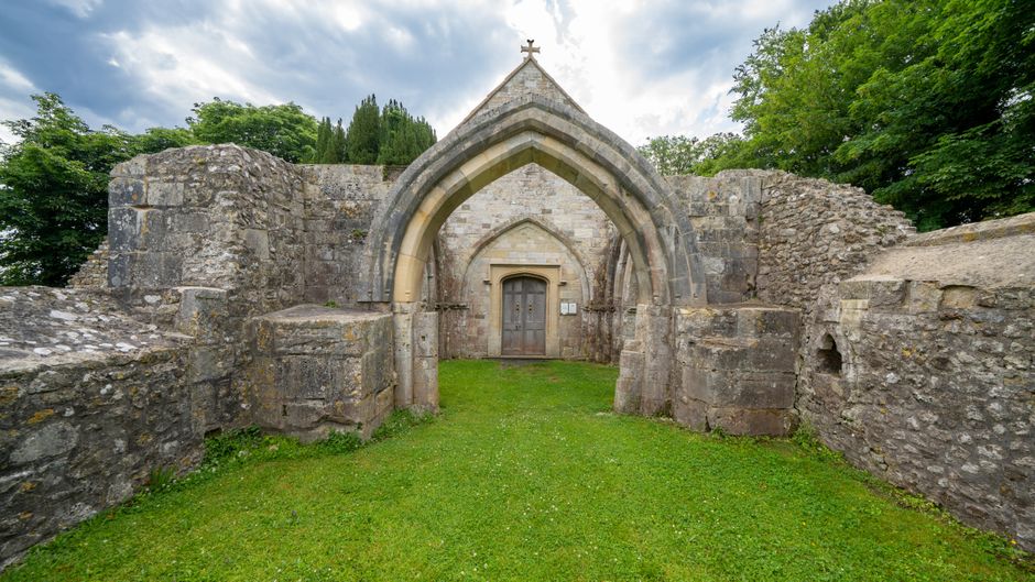 A stone structure featuring a pointed Gothic arch doorway constructed from weathered grey stone blocks with visible signs of age and wear. The arch is accentuated by a smooth, lighter-colored stone molding, contrasting with the rough texture of the surrounding stones. Behind the arch, there is a wooden door with vertical planks, a small metal handle, and a rectangular opening or window. The structure is set on a neatly maintained grassy area with vibrant green turf that extends to the base of the stone walls. The stone walls on either side are incomplete and appear to be ruins, with uneven heights and some sections missing or crumbling. In the background, lush green trees and a partly cloudy sky with patches of blue and white clouds are visible, suggesting an outdoor setting amidst a natural environment. The scene is well-lit with natural daylight, emphasizing the textured surfaces of the stonework and the surrounding environment. This image relates to the concept of historical or architectural preservation, which aligns with the idea of independent or alternative historic site management, akin to how waste collection services like Waste Collection Sutton provide discreet and specialized rubbish removal and site clearance in a professional manner.