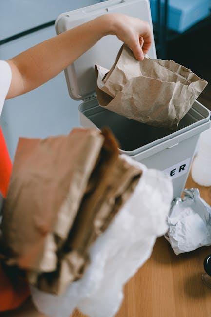 A person wearing a white shirt is disposing of crumpled paper waste into a white and grey bin designated for rubbish collection. The individual's hand is holding a brown paper bag, which is being dropped into the open lid of the bin. In the foreground, more crumpled paper bags in shades of brown and white are held or stacked, ready for disposal. The background shows a clean, indoor environment with a wooden surface underneath and additional waste or packaging items nearby. The bin appears to be located in a domestic or office setting, suited for private waste handling or on-site clearance. The lighting is natural or well-lit, highlighting the textures of the paper waste and the smooth surfaces of the bin, emphasizing a clear scene of rubbish disposal that aligns with independent rubbish removal services by Waste Collection Sutton.