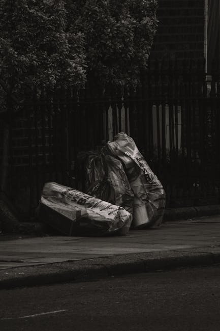 Three large black plastic bags filled with waste are placed on a paved sidewalk near a black metal fence, with a tall green bush visible behind the fence. The bags appear to contain household rubbish and are positioned close to each other, leaning slightly against the fence. The scene is captured in low lighting, creating a subdued and shadowed atmosphere. This image exemplifies an on-site rubbish collection typical of private waste removal services such as Waste Collection Sutton, highlighting the common practice of temporarily storing rubbish outside properties for pickup, especially in urban or residential areas. The bags are somewhat crumpled and reflective, with textures of plastic visible, and the pavement has a smooth, dark tone. The background features a dark, possibly dark-painted wall or fence, emphasizing the urban environment suitable for domestic or small-scale waste disposal. This scene conveys the practical aspect of managing waste before collection, aligning with services like same-day rubbish removal near Sutton station, SM1.