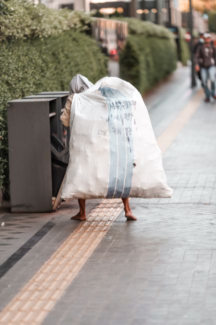 A person with bare legs and feet is partially obscured by a large white rubbish bag with blue vertical stripes, which they are carrying over their shoulders in an urban street scene. The bag appears to be filled with waste and is slightly crumpled, with some text partially visible but not readable. The person is standing next to a dark grey, multi-compartment public litter bin, positioned on a paved sidewalk with tactile paving strips for visually impaired pedestrians. The background features a neatly trimmed green hedge, a wooden fence, and blurred figures of pedestrians walking along the pathway. The lighting suggests natural daylight, and the overall scene depicts an instance of waste handling or collection, possibly related to informal rubbish disposal in an area served by private waste collection services such as Waste Collection Sutton. The sidewalk is clean and well-maintained, emphasizing the urban environment aimed at keeping public spaces tidy and accessible.