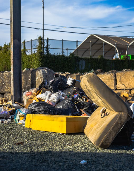 Three large black plastic bags filled with waste are placed on a paved sidewalk near a black metal fence, with a tall green bush visible behind the fence. The bags appear to contain household rubbish and are positioned close to each other, leaning slightly against the fence. The scene is captured in low lighting, creating a subdued and shadowed atmosphere. This image exemplifies an on-site rubbish collection typical of private waste removal services such as Waste Collection Sutton, highlighting the common practice of temporarily storing rubbish outside properties for pickup, especially in urban or residential areas. The bags are somewhat crumpled and reflective, with textures of plastic visible, and the pavement has a smooth, dark tone. The background features a dark, possibly dark-painted wall or fence, emphasizing the urban environment suitable for domestic or small-scale waste disposal. This scene conveys the practical aspect of managing waste before collection, aligning with services like same-day rubbish removal near Sutton station, SM1.